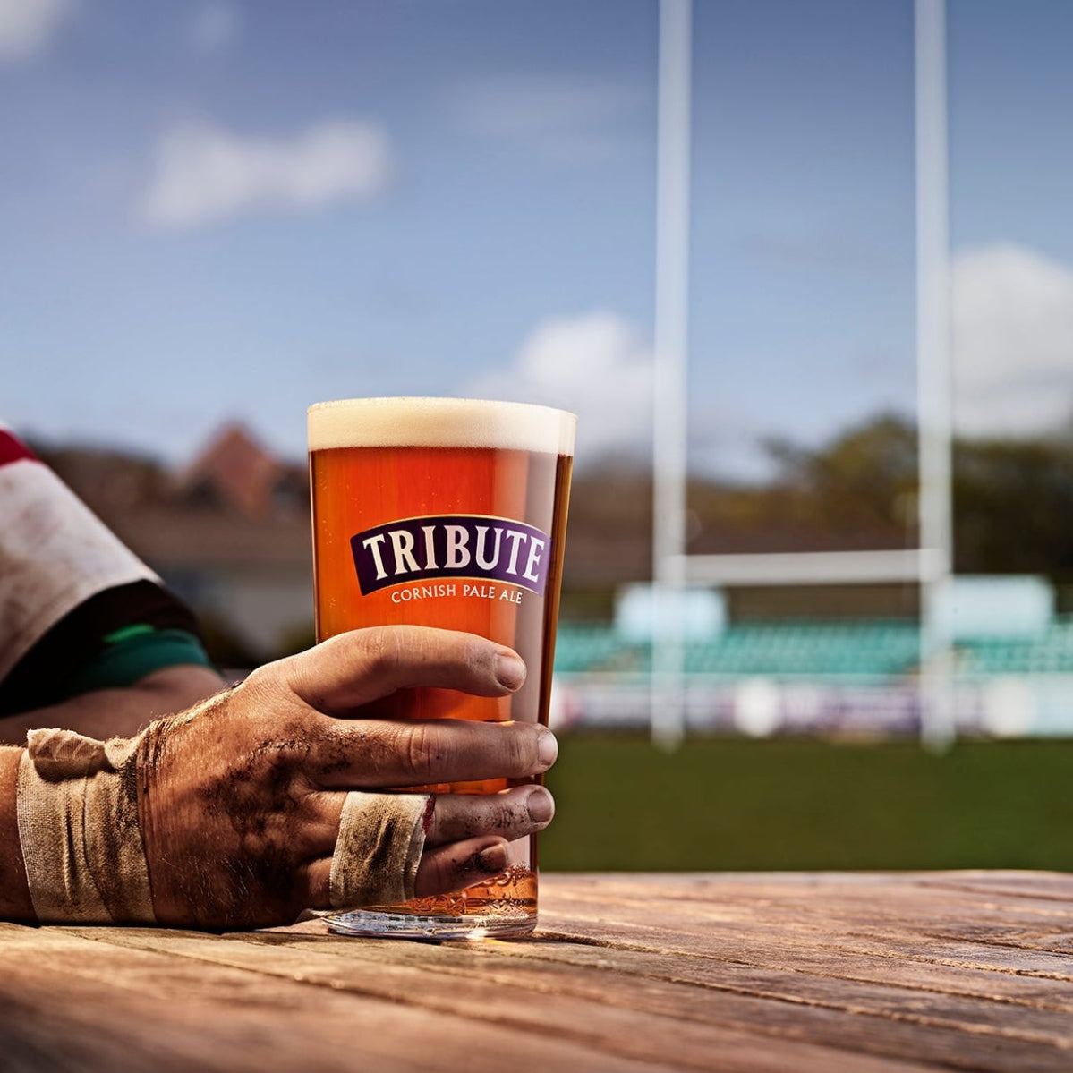 Hand holding a glass of Tribute beer at a rugby field with goalposts in the background